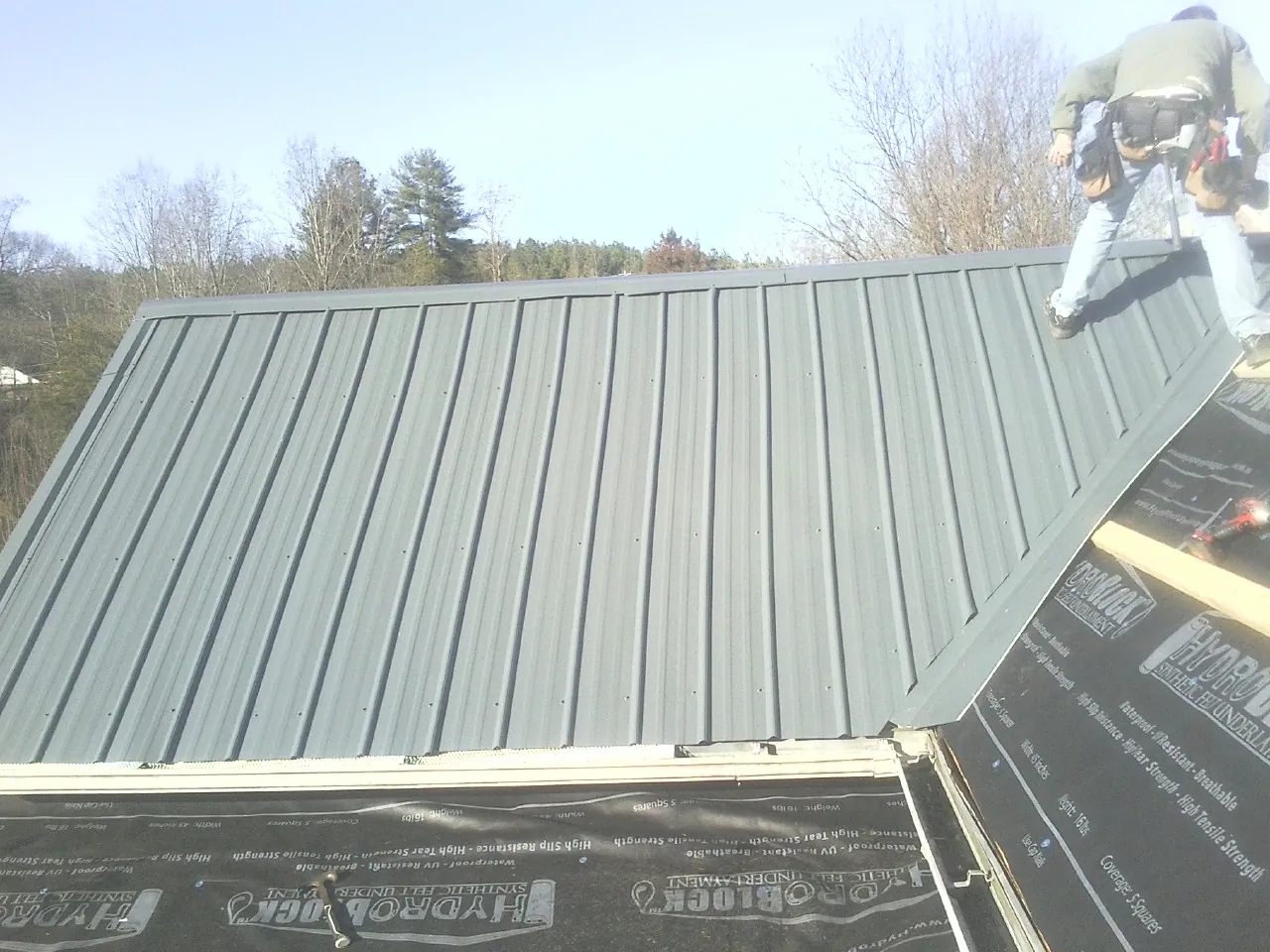 Crew installing dark green standing seam panels over synthetic underlayment in Perris