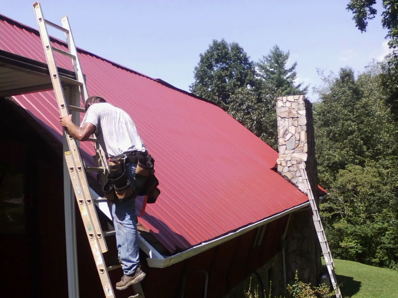 Roofer on a ladder beside a red metal roof and stone chimney — Soffit & Fascia Repair by Roofing Brothers in Perris